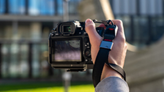 Wir sehen eine Hand und eine Kamera in der Hand. Mediamatiker bei der Arbeit.